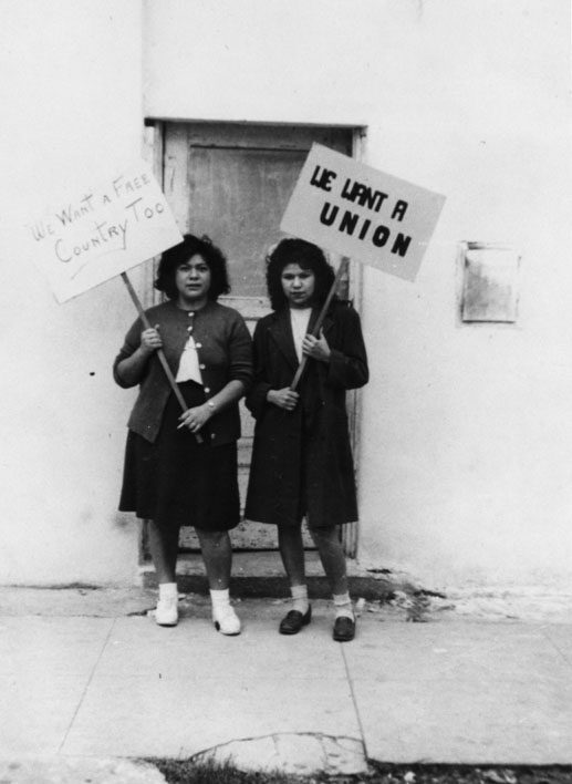 Two Mexican-American women holding picket signs outside a factory. The woman on the right's says "We Want a Union" and the woman on the left's says "And a Free Country Too." Both wear pleated skirts, sweaters, and bobby socks typical of the 1940s.