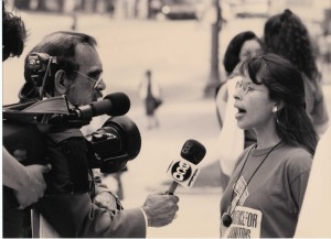 black and white image of a young woman being interviewed by a newscaster and his camera man. The newscaster wears a suit and holds a microphone labelled "ECO." The young woman speaking wears glasses, a Justice for Janitors t-shirt, and a necklace.