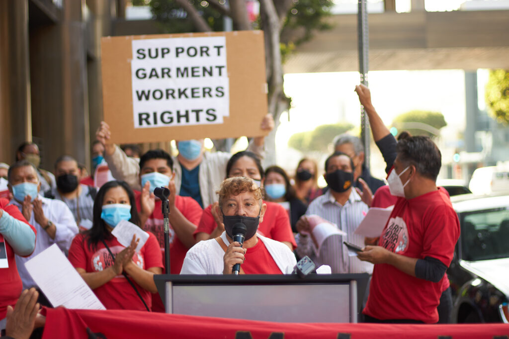 An older woman wearing a face mask addresses a crowd at a podium. She is wearing a red t-shirt, as are many in the crowd behind her. One member of the cheering crowd holds a sign that reads "Support Garment Worker Rights". Some raise their fists.