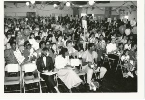 Janitorial workers, some with their children, gather in a hall decorated with streamers and balloons. Rows of chairs are full with smiling workers. Some have signs on the ends indicating various delegations and shop committees.