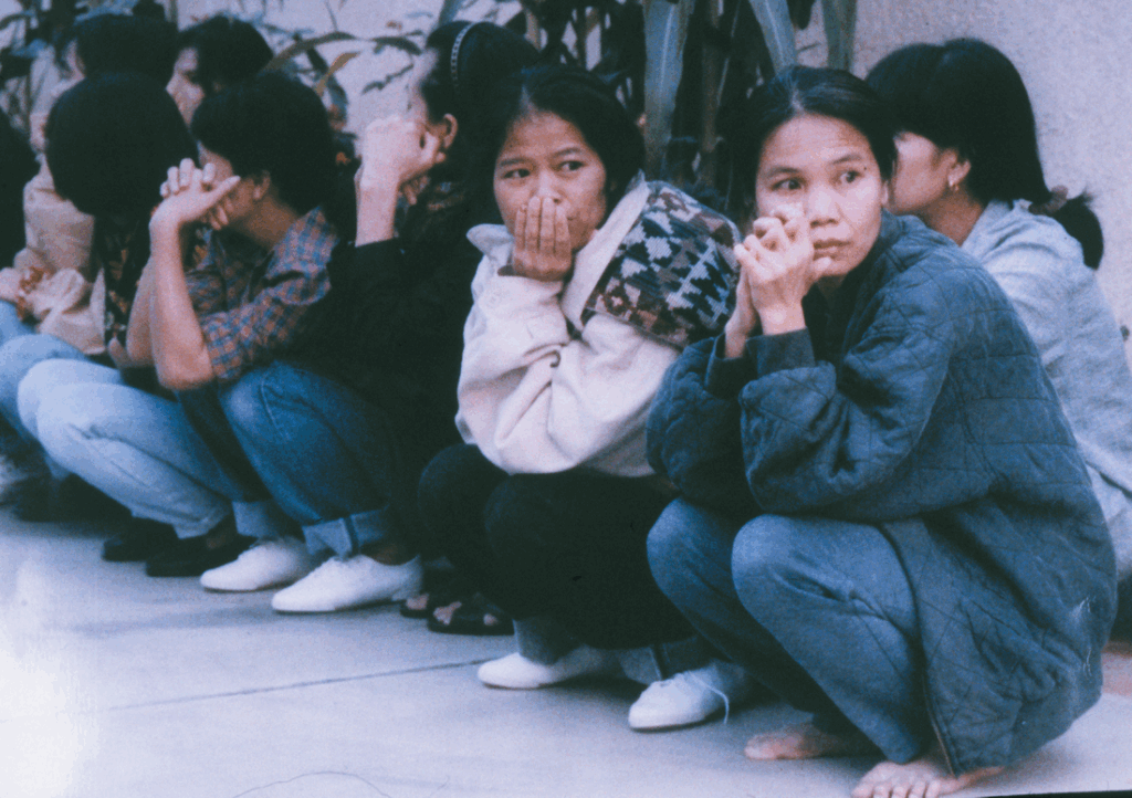 A group of Thai women wearing winter clothes are crouching, many with their hands gathered near their faces. Only two in the foreground of the image look towards the camera, both of whom appear to have been crying and look very concerned