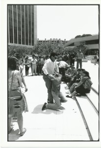 black and white photo of an outdoor civil disobedience training. In the center of the image, two men mimic an arrest, which one man taking the other into custody and dragging him away. surrounding them a crowd watches on.