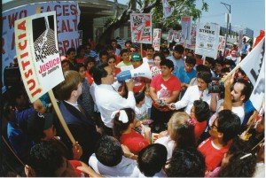 color image with stereoscope effect shows a group of janitors gathered around a man attempting to address their concerns on a megaphone. some carry picket signs reading "L.A. Should Work for Everyone" and "Justice for Janitors." A banner hangs on the side of the building, partial obscured, reading "L.A.'s Two Faces: Glamour and Wealth, Poverty and Despair"