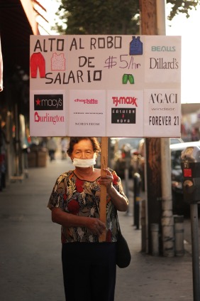 Striking image of an older woman holding a handmade sign on a street corner with the sun setting behind her. Her sign reads "Alto Al Robo de Salario" and bears the corporate logos of major department stores including Macys, Burlington, and Dillards.