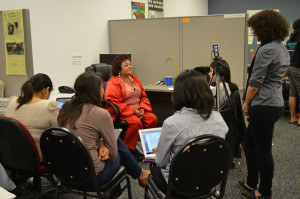 an older Latina in an all red outfit sits in a cubicle of an office. She is surrounded by a group of students (their backs to the camera) interviewing her. One student stands recording the interview on a phone.