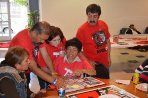 A group of five Latino-presenting people browsing through a photo album and talking. Four of them wear read Justice for Janitors t-shirts.