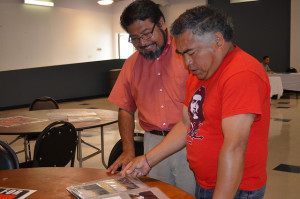 Photograph of two Latino men, one older in a red Che t-shirt flipping through a photo album, the other in a short sleeved button up shirt smiling while listening to him.