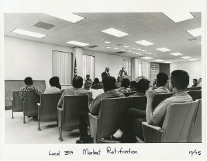 black and white photograph of union members sitting in a union hall, their backs to the camera, listening to a speaker at the podium.
