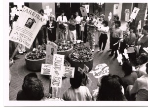 A group of people holding signs gather in a circle around four large planters in a courtyard. Some hold classes reading "Jobs with justice," some dove shaped signs reading "We Want justice" and others picket lines reading "Arriba - Justicia Para Los Janitors."