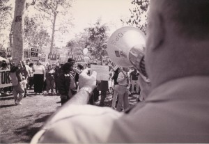 Black and white image taken over the shoulder of a police officer speaking into a megaphone as people march carrying picket signs.