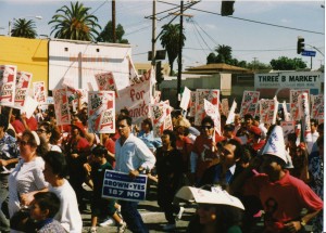 scene of a protest march on Cesar E. Chavez Ave. in Boyle Heights (the street sign visible in the background). Protestors carry signs including "Brown Yes - 187 No" and "L.A. Should Work for Everyone"