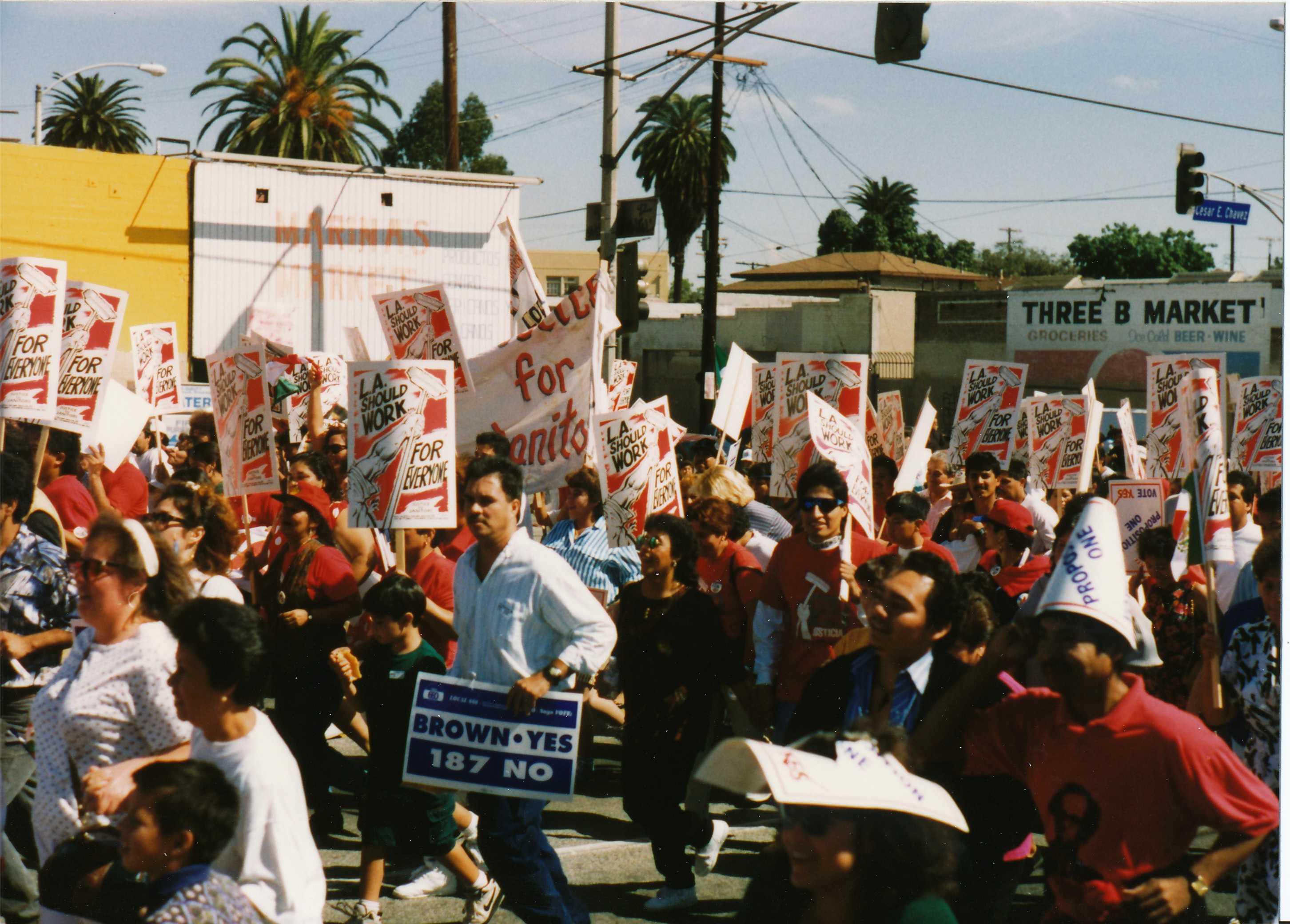 scene of a protest march on Cesar E. Chavez Ave. in Boyle Heights (the street sign visible in the background). Protestors carry signs including "Brown Yes - 187 No" and "L.A. Should Work for Everyone"