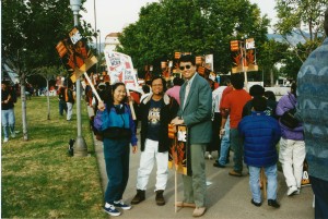 Three Asian American holding picket signs at a demonstration. The tall man on the right wears sunglasses and a suit and tie. They carry pickets reading "April One" and "L.A. Should Work for Everyone"