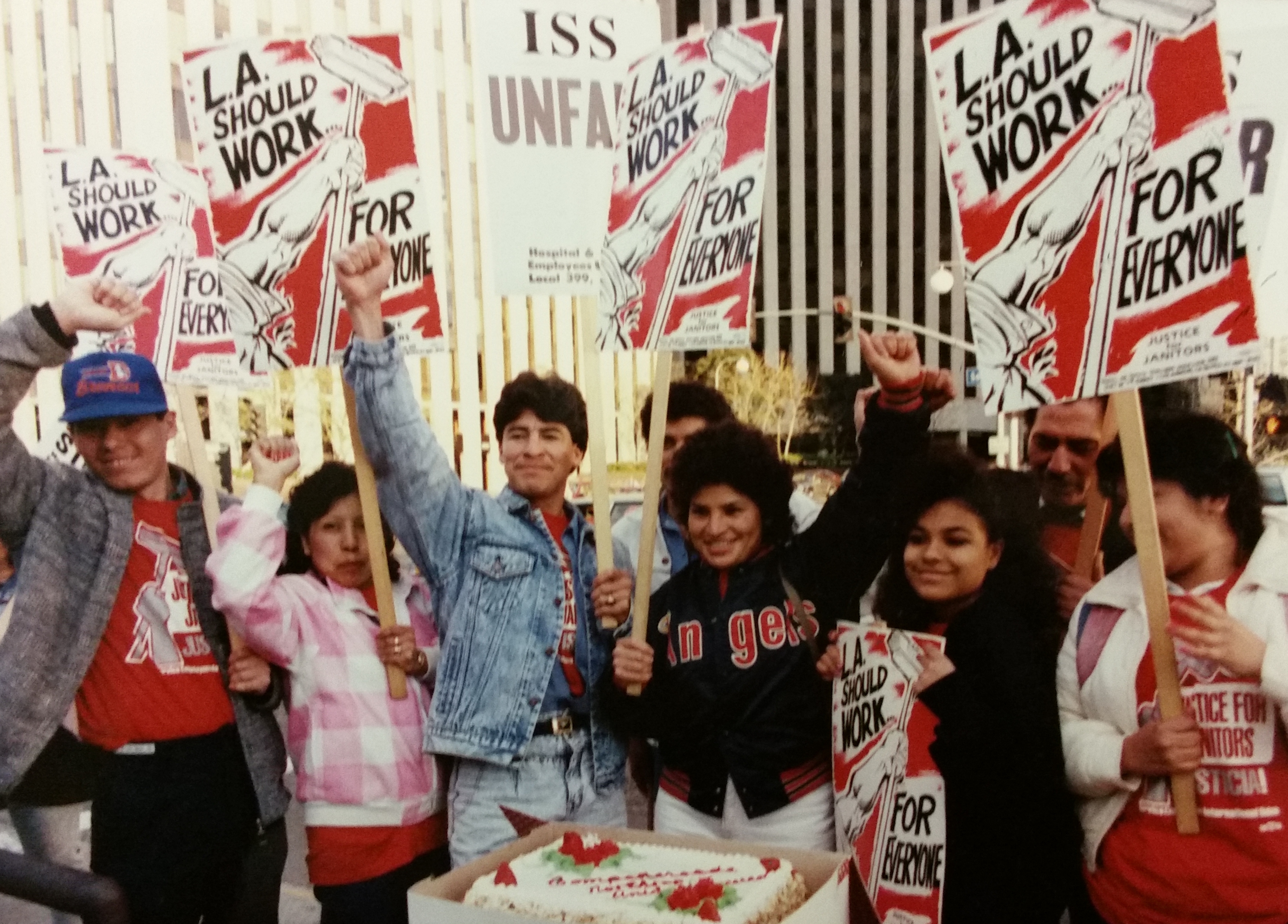 Color photo of a group of smiling janitors with their fists raised holding red, white, and black picket signs reading "L.A. Should Work for Everyone" with a graphic of a raised fist holding a mop. Several are wearing red Justice for Janitors t-shirts.