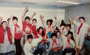 A group of janitors gathered in a locker room smiling and raising their fists. Several are wearing gray workshirts with sewn-on nametags. Most of the group is wearing red Justice for Janitors t-shirts.