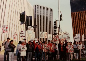 Color photo of a group of smiling janitors with their fists raised holding red, white, and black picket signs reading "L.A. Should Work for Everyone" with a graphic of a raised fist holding a mop. Several are wearing red Justice for Janitors t-shirts. Behind them loom large glittering office towers.