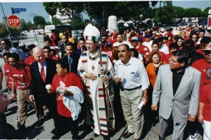 Striking color image from a protest on Spring Street in downtown Los Angeles. In the center of the image is an Archbishop wearing his vestments and carrying a large staff. In striking symmetry, one of the janitors behind hi is carrying a mop at almost the exact same angle.