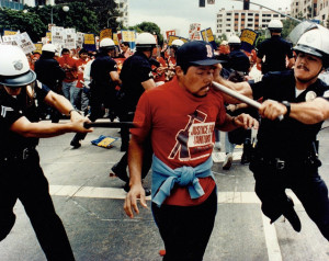two police officer swing their batons into body of a janitor, each hitting him from one side. The man in the middle is wearing a red union t-shirt and a Red sox hat, his face distorted as one of the batons hits his jaw.