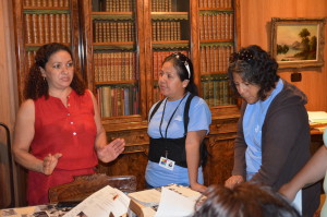 Three Latinx woman in a fancy library room, with rare books displayed in a glass cabinet behind them and a painting on the wall. One woman is speaking, another watching her as she speaks, and the third is looking through the archival documents displayed on the table in front of them.