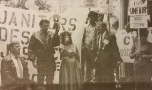 Black and white image of speakers at a rally standing in front of a large banner reading "Janitors Demand Justice." One man holds a megaphone while a woman in the center speaks into it. the workers surrounding them hold picket signs reading "ISS UNFAIR"