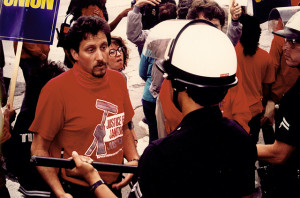 A young man with a goatee and a scarf around his neck speaks with a police officer in a helmet, his back facing the camera. The man wears a red Justice for Janitors t-shirt.