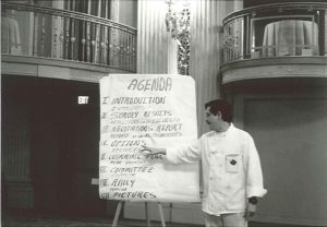 Black and white image of a man in a chef's coat in what appears to be an elegant ballroom with balconies above. The man is pointing to a large piece of paper on a easel that says "AGENDA" and lists Introduction; Survey Results; Negotiations Report; Options; Winning Plan; Committee; Rally; Pictures - all written in Spanish and English.