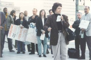 A woman in a sport coat speaks at an outdoor rally on a small microphone. People behind her hold signs. The street lamp in the background reads "1st St."