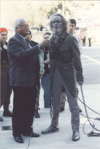 On the right, Dave Clennon dressed in a full costume, complete with chains and make up, addressing the media as Jacob Marley on his way to address the City Council. Rev. James Lawson holds the microphone for him.