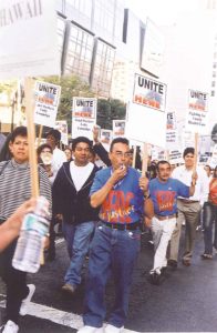 workers marching carrying picket signs reading "UNITE HERE". some wear blue UNITE-HERE t-shirts.