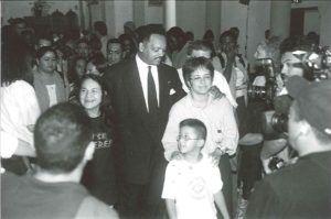 Black and white image of Rev. Jesse Jackson, wearing a suit, walking with his arms around two women: Dolores Huerta (to his right) and Maria Elena Durazo (to his left). Huerta wears a shirt that says Is Se Puede. Durazo, who looks tired, walks with her young son Michael.