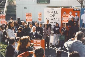 Scene of an outdoor press conference in a courtyard near Little Tokyo in downtown Los Angeles. Reporters carrying cameras stand in the foreground with their backs turned to the camera. At the podium is an older Asian man with long white hair tied back. There is a crowd of people behind him, some holding picket signs reading "New Otani - Non-Union - please Boycott!" On the right hand sign of the frame is a larger free-standing poster titled "Wall of Shame" with graphic images of starved detainees.
