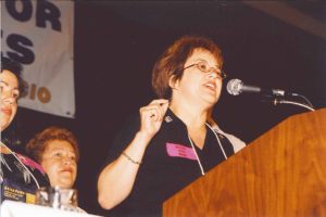 A woman with short hair and glasses speaks at a podium.