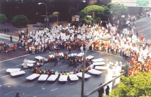 Photograph taken from above an intersection where a civil disobedience action is taking place. At the center of the image, people sit in a circle cross legged, preparing to be arrested. A priest in his vestments blesses their action. A ring of hotel beds encircles them, and a few yards beyond that, a crowd of people with signs and banners.