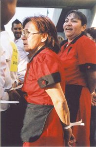 Two women in red and black maid's uniforms with their hands cuffed behind their backs. The woman in the forefront looks vexed, but the woman behind her is laughing.