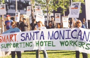 Two older woman and a teenage girl hold a large banner reading "S.M.A.R.T. Santa Monica's Supporting Hotel Workers." They are smiling. Behind them, people hold UNITE HERE picket signs.