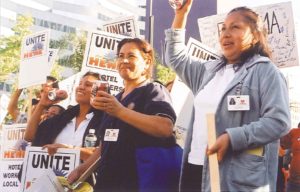 three women shake noise makers made from coca cola cans at a picket line. The signs surrounding them read "UNITE HERE." One woman wears her uniform and two wear bases from their worksite.