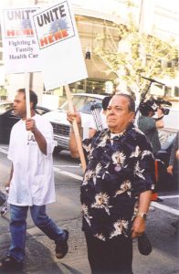 An older man in a Hawaiian print t-shirt stands holding a UNITE HERE picket sign.