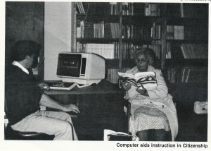 A black and white image of a photograph cropped from a newsletter. The caption reads "Computer aids instruction in citizenship" and it shows an older woman sitting in a chair in a library reading a book with "citizenship" on the cover. A man at a desk assists her as he works on an ancient looking computer