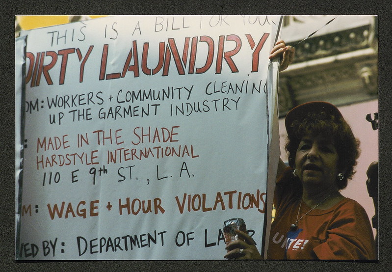 An older woman in a UNITE! t-shirt holds a big sign that reads: "This is a bill for your DIRTY LAUNDRY from: Workers and Community Cleaning up the Garment Industry -- to: Made in the Shade hardstyle International, 110 E. 9th St., L.A. - Wage and hour Violations - by: Department of Labor." In her hand is a soda can sealed with duck tape, likely a home made noise maker.