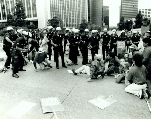 Black and white image of a cop riot in process. On the left hand side of the image, officers brandishing billy clubs are lined up, ready to strike. one officer raises his hand barking orders, while others swing their clubs at men on the ground. On the right, a crowd of janitors in distress, many of them on the ground, brace themselves. Their pickets are strewn in the street.