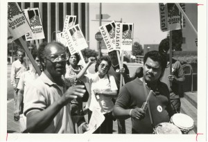 black and white image from a picket line downtown Los Angeles. in the foreground, an African American man directs the marchers. A man with a 80s style mullet plays a drum