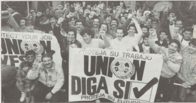 Black and white image featuring a group of men raise their fists in celebration. The hold a sign in Spanish reading, "Union, Yes!"