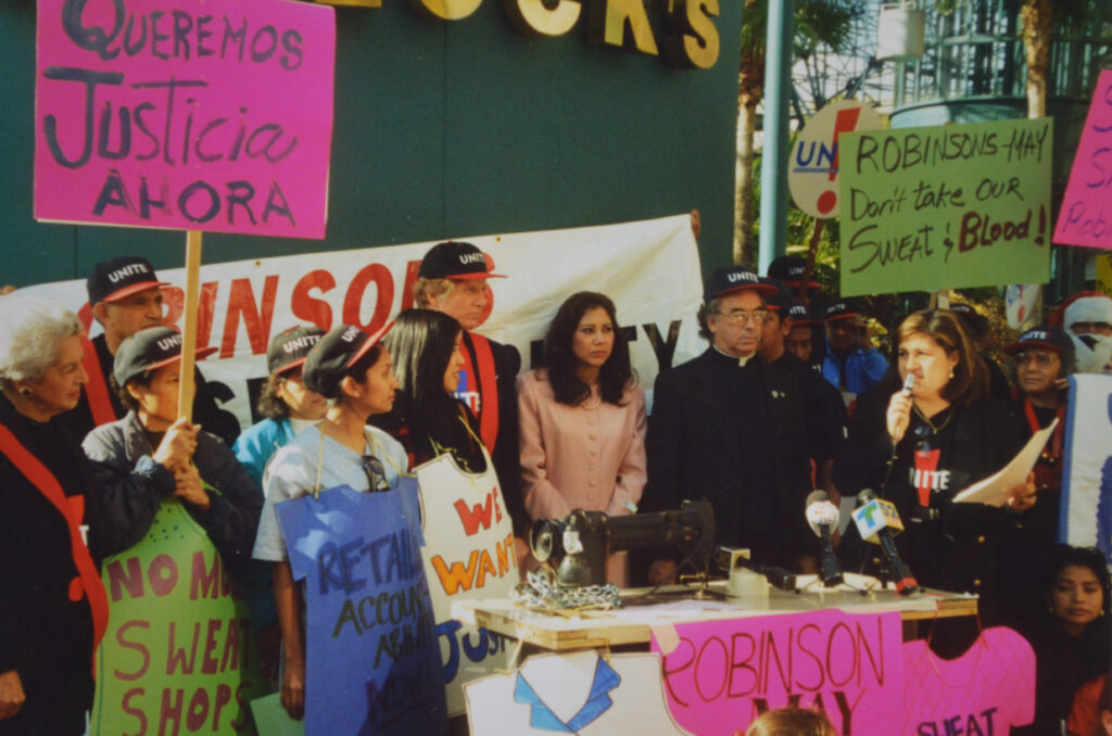 a proud of people, many of them wearing black and red UNITE! hats and/or carrying handmade picket signs, gather outside a store in Santa Monica. On the right, a young speaks its a microphone.