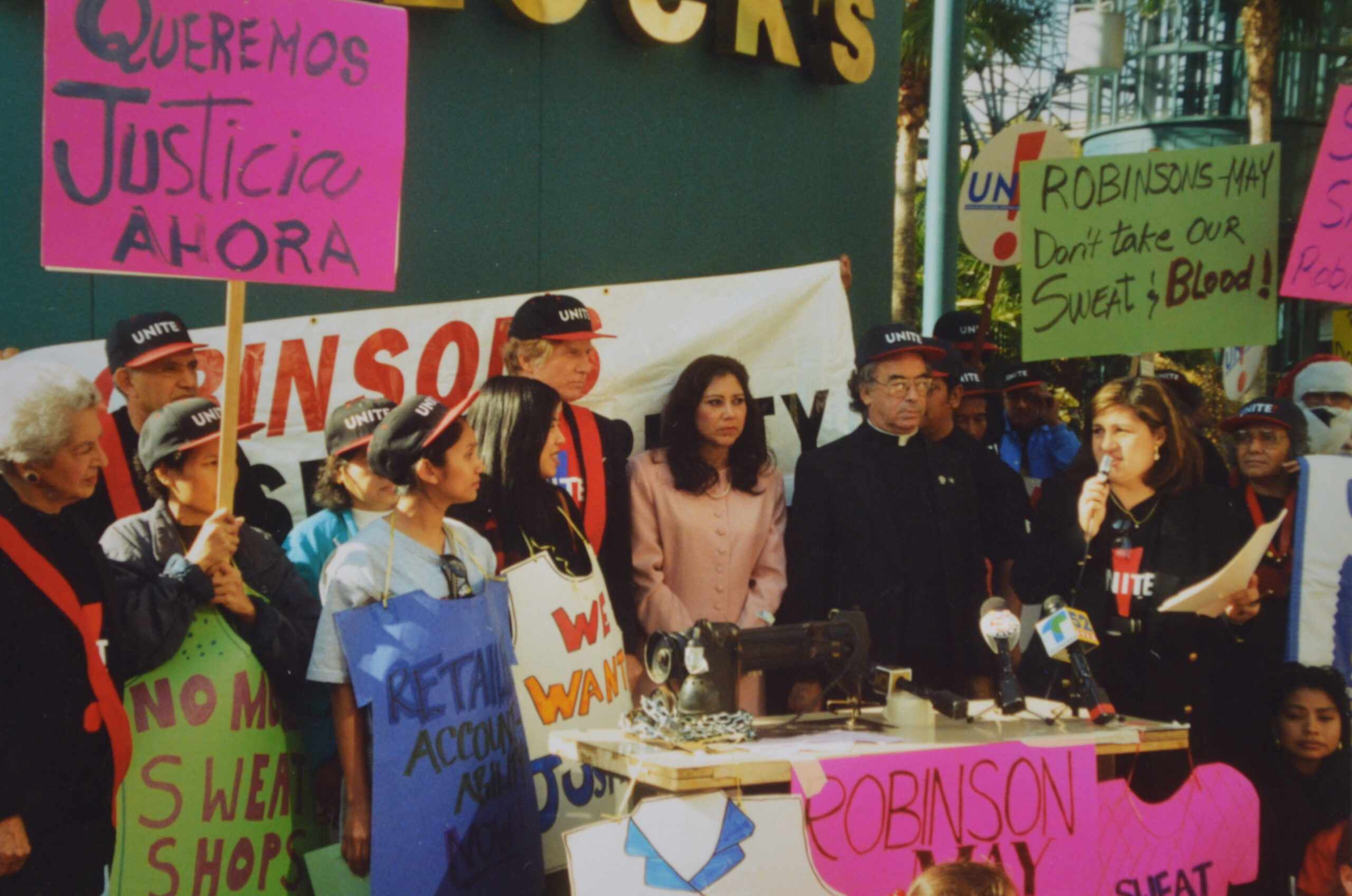 a proud of people, many of them wearing black and red UNITE! hats and/or carrying handmade picket signs, gather outside a store in Santa Monica. On the right, a young speaks its a microphone.