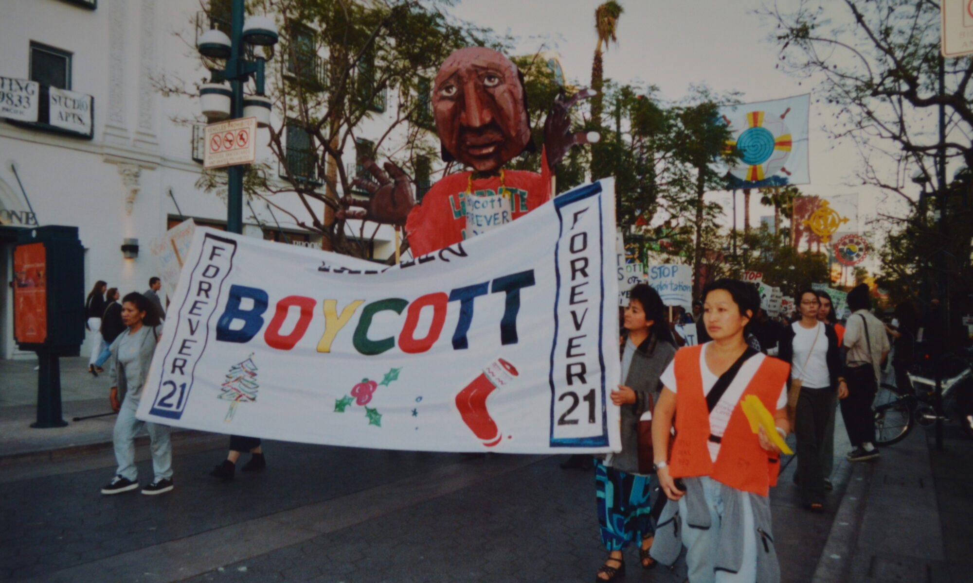Image from a demonstration on the Santa Monica 3rd street promenade. Two protestors hold a larger banner reading "Boycott" with Christmas holiday symbols. Behind them looms a large, handmade puppet being held up by other protestors. On the right side of the march, a woman directing the protestors wears an orange safety vest.