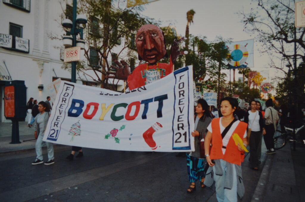 Scene from a demonstration at the Third Street Promenade shows workers carrying a large banner reading "BOYCOTT" and a paper mache puppet walking behind them.