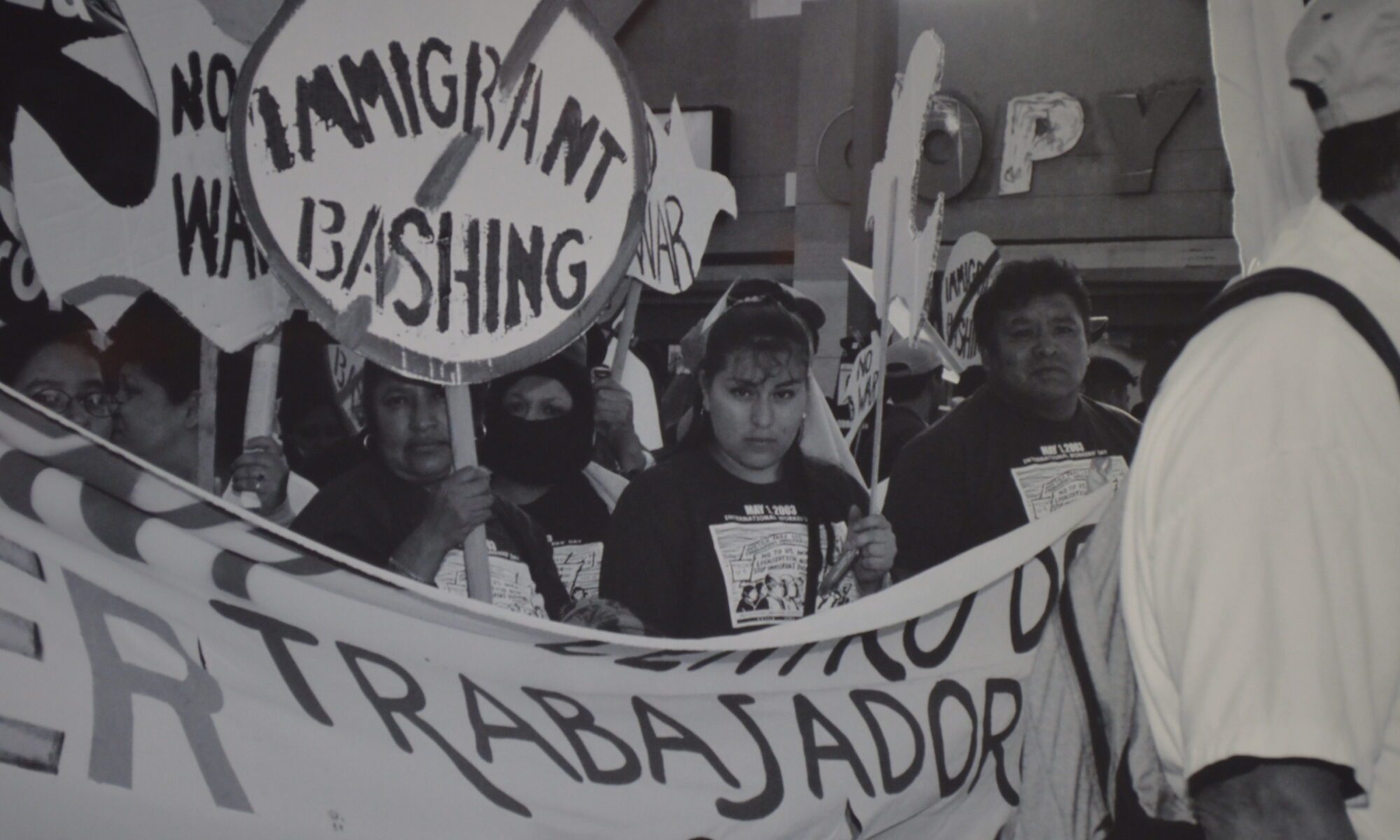 Black and white image taken at a May Day march. Two garment workers are carrying a large banner from the Garment Workers Center, the only word fully visible is "Trabajador." other people in the image are carrying picket signs, one that says "immigrant bashing" that is crossed over. and another is shaped like a dove and reads "no war"