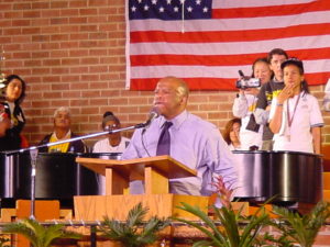 Congressman John lewis, his face glistening with sweat, addresses an audience at what appears to be the lecturn in a church. Behind him is a large American flag.
