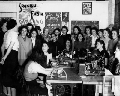 Black and white image of a classroom full of women, some seated at sewing machines, others standing. Signs on the back wall read "Spanish Fiesta- Dancing" and "Sign up now - New Mandolin Class"