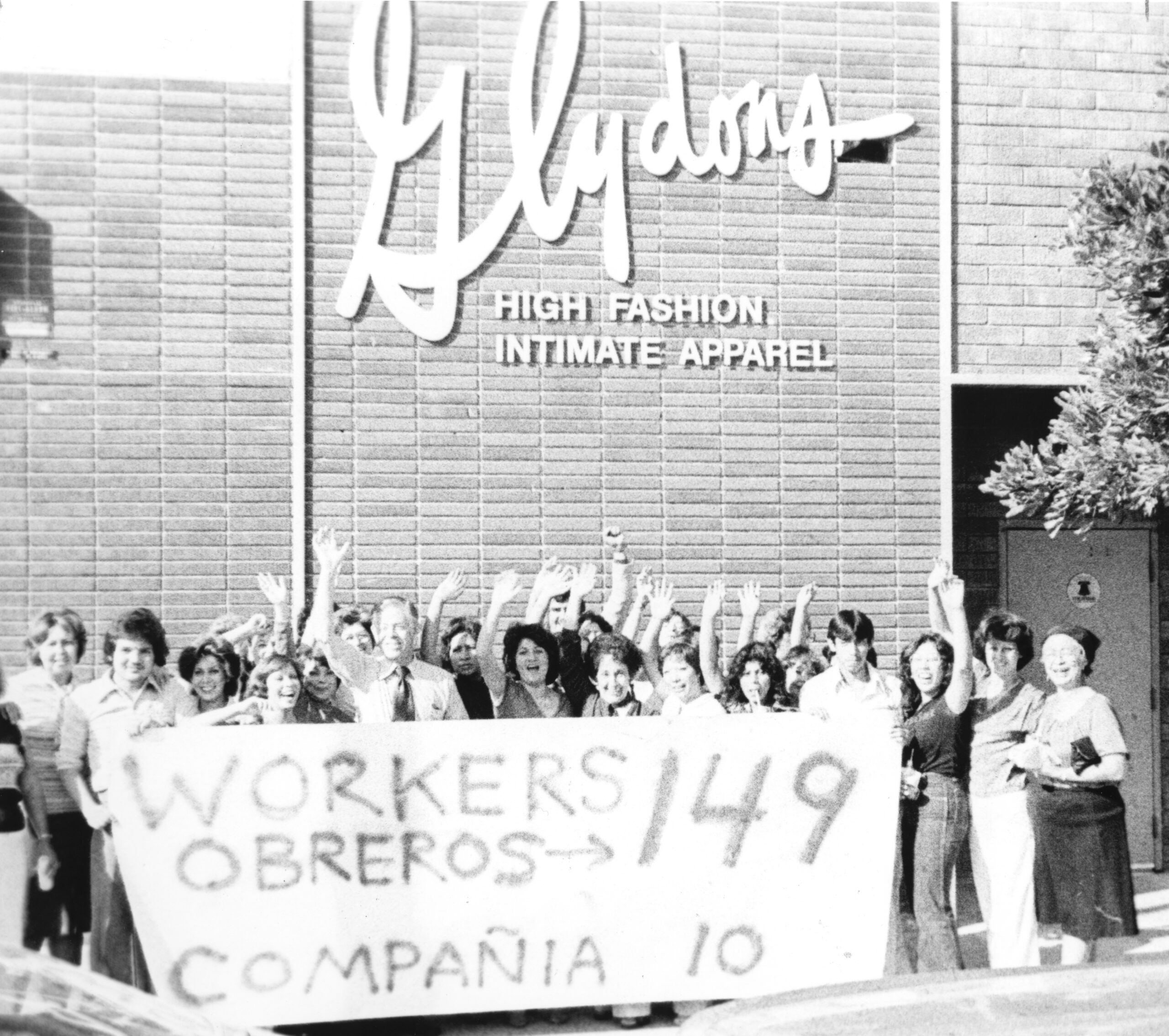 Workers celebrate a union victory outside of a garment factory in Los Angeles, 1980. They stand waving and smiling while holding a sign that indicates that 149 workers voted for the union and 10 against it.